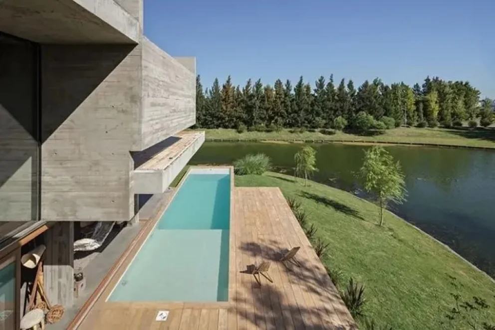 Espacio exterior del hogar con una pileta de gran magnitud, piso de madera y un quincho semiabierto, al frente de la laguna